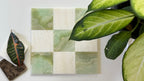 Checkered tile pattern with green and beige squares on a white surface, surrounded by leaves.